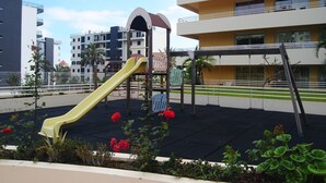 A playground with a yellow slide and swings, surrounded by a garden with red flowers, in an urban setting with multi-story buildings in the background. - Aurora Park with swimming pool (Funchal)