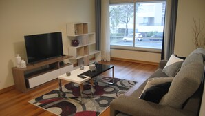 A modern living room with a flat-screen TV, a coffee table, a sofa, and a large window offering a view of the street. - Aurora Park with swimming pool (Funchal)
