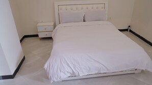 A neatly made bed with white bedding in a room with white walls and a white nightstand with drawers.