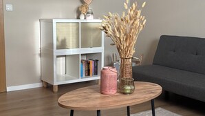 Un salón con una mesa de centro redonda de madera, un mueble blanco con libros, un sofá gris y un televisor de pantalla plana montado en la pared.