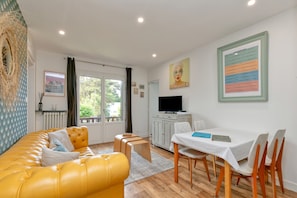 A living room with a yellow sofa, a dining table with chairs, a television, and artwork on the walls.