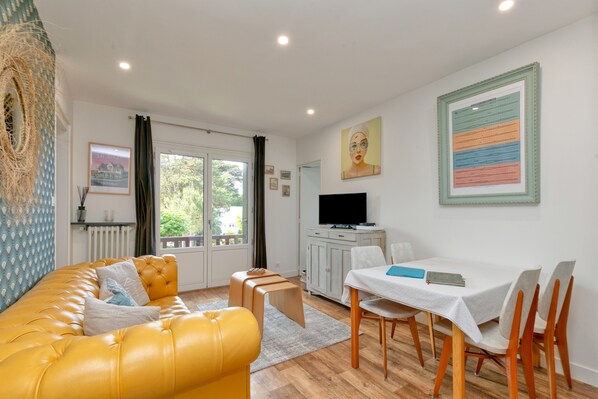A living room with a yellow sofa, a dining table with chairs, a television, and artwork on the walls.