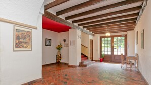 Un couloir au sol recouvert de briques rouges, avec des poutres en bois, un escalier et une porte donnant sur l’extérieur.