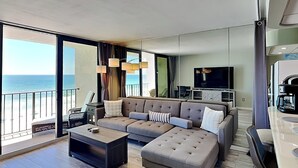 Un salon moderne avec un grand canapé, une table basse et un téléviseur. La pièce donne sur un balcon offrant une vue sur l’océan.