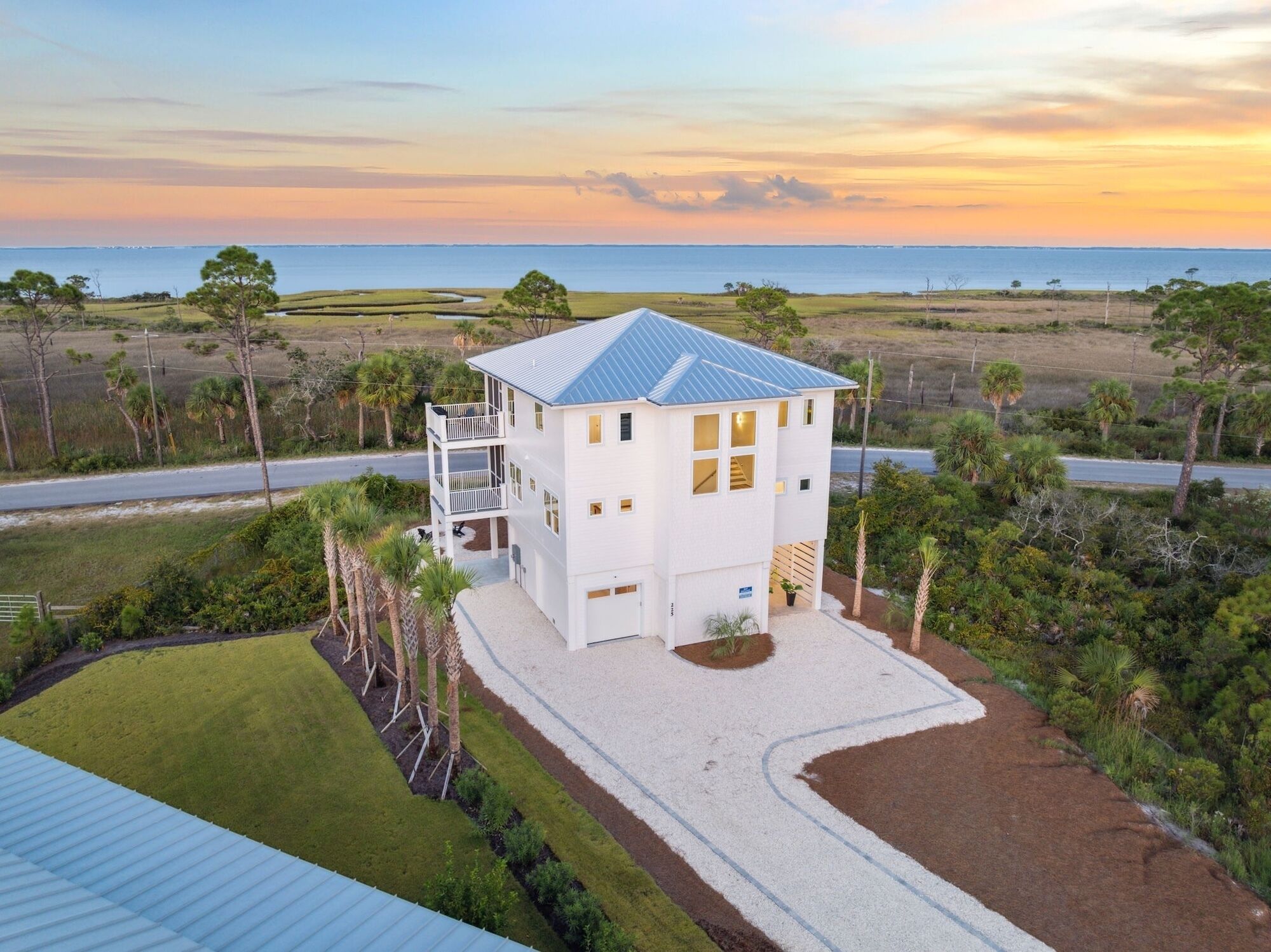 A white two-story house with a blue roof, surrounded by greenery and a view of the ocean at sunset.