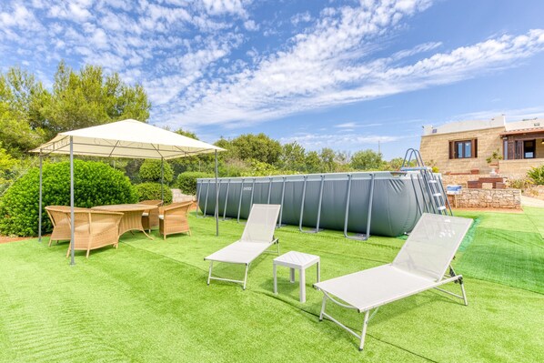 Un jardin avec une pelouse artificielle, une piscine, des chaises longues, une table et un espace abrité.