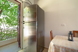 Una habitación compacta con un refrigerador de acero inoxidable, una mesa con mantel a cuadros, un farol y un balcón con barandilla de metal y árboles.