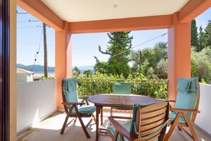 Un espacio de estar al aire libre techado, con una mesa y sillas de madera, vistas a zonas verdes y montañas, y una pared blanca.