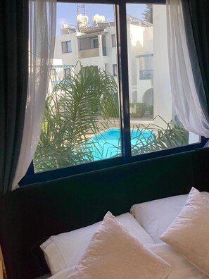 Camera d'albergo con un letto, cuscini bianchi e vista su una piscina e edifici esterni.