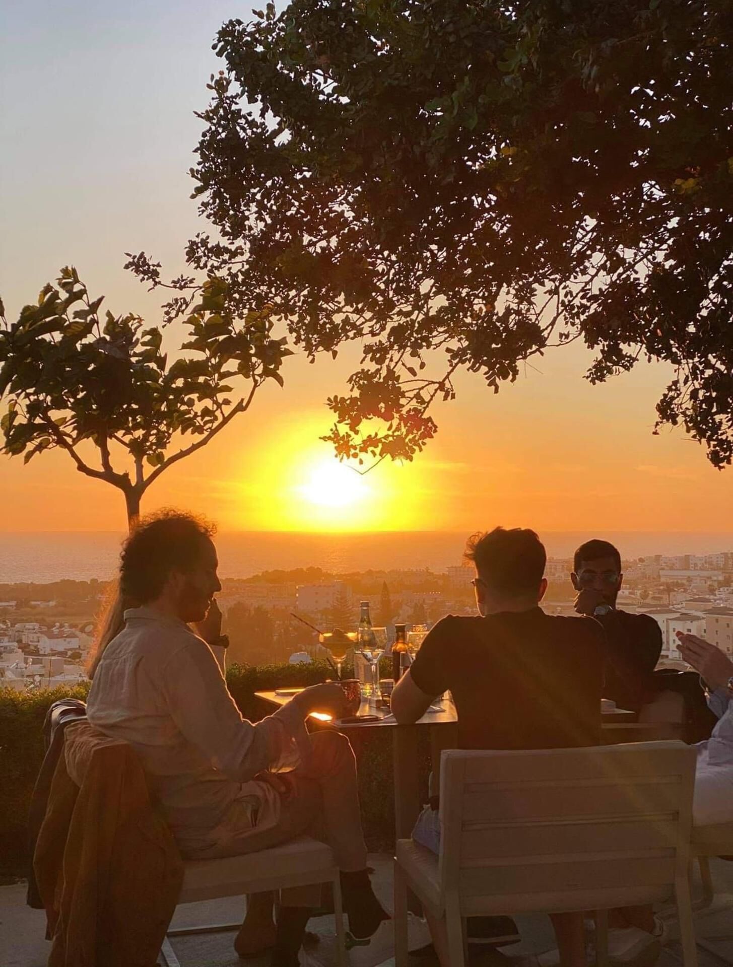 Un grupo de personas sentadas en una mesa al aire libre con vistas a la puesta de sol sobre un paisaje urbano.