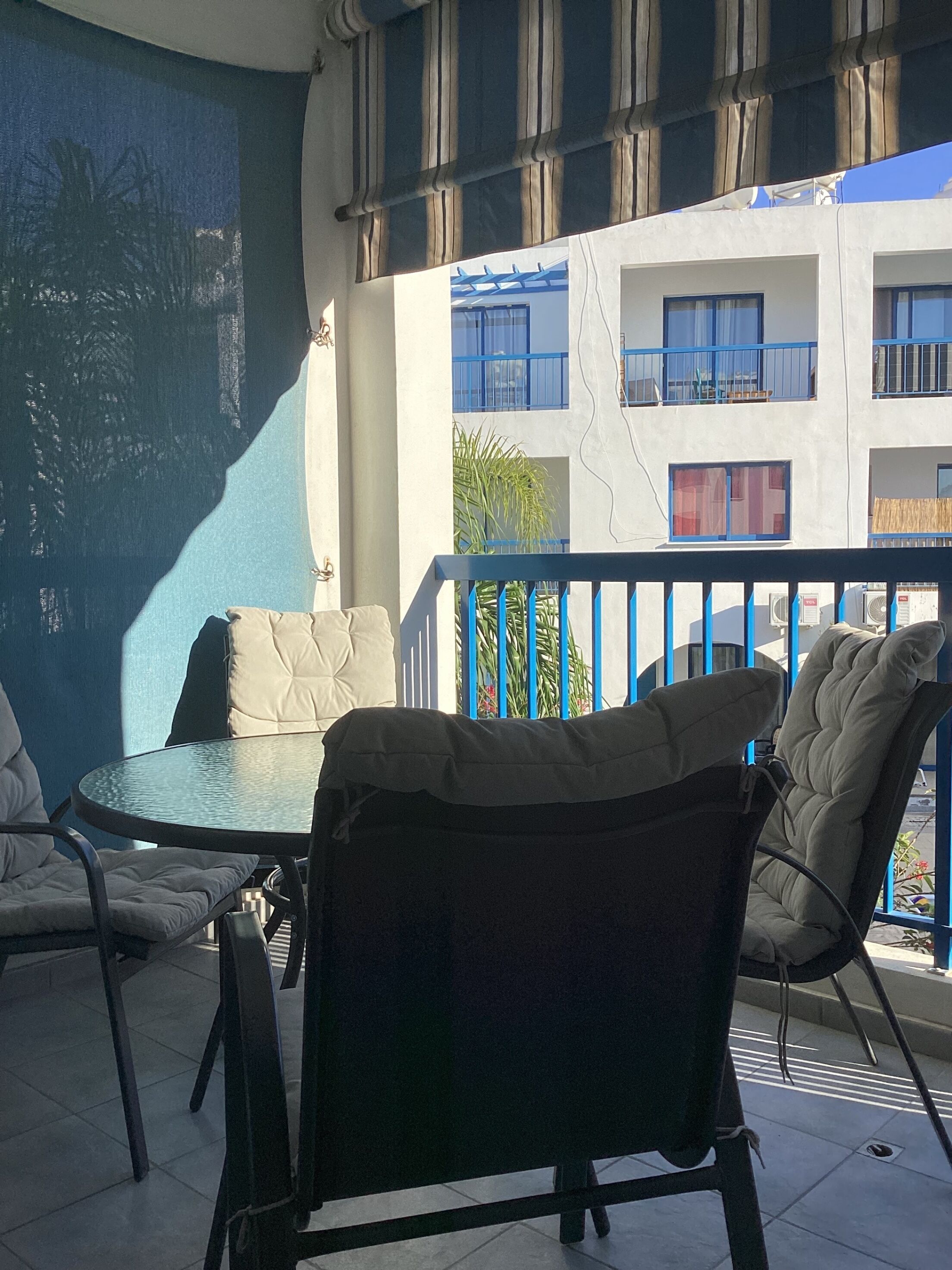 A balcony with a glass table and chairs, a view of a building with blue railings, and a blue wall.