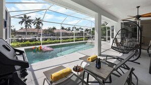 A poolside area with a glass enclosure, outdoor seating, a grill, and a ceiling fan. - Seaside Waterfront Home | Heated Pool & Dock (Marco Island)