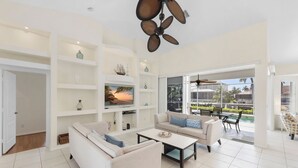 A spacious living room with a large TV, white sofas, a coffee table, and a ceiling fan. - Seaside Waterfront Home | Heated Pool & Dock (Marco Island)