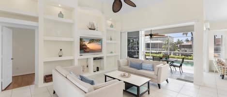 A spacious living room with a large TV, white sofas, a coffee table, and a ceiling fan.