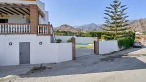 Una propiedad residencial con fachada blanca, un balcón, una antena parabólica, un alto árbol de hoja perenne y montañas al fondo.
