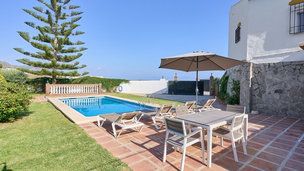 Una piscina con sillones de descanso y una sombrilla, un muro de piedra y un árbol grande.