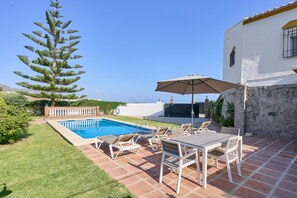 Una piscina con sillones de descanso y una sombrilla, un muro de piedra y un árbol grande.