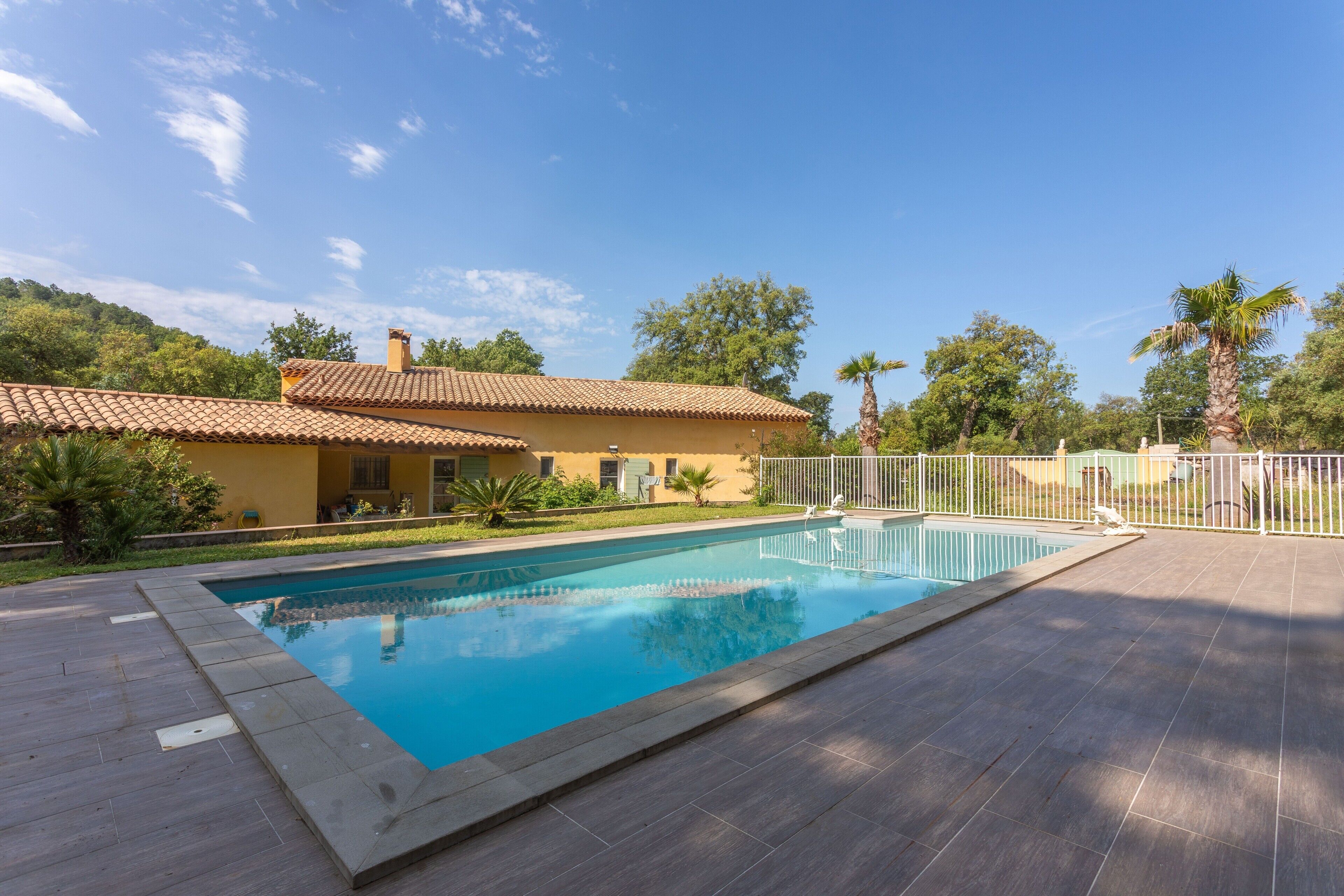 A swimming pool with a tiled deck, a yellow building with a tiled roof, and a fenced area with palm trees.
