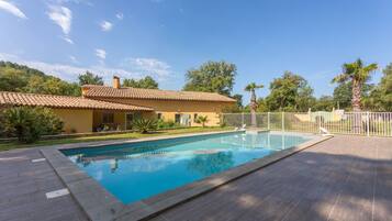 A swimming pool with a tiled deck, a yellow building with a tiled roof, and a fenced area with palm trees.