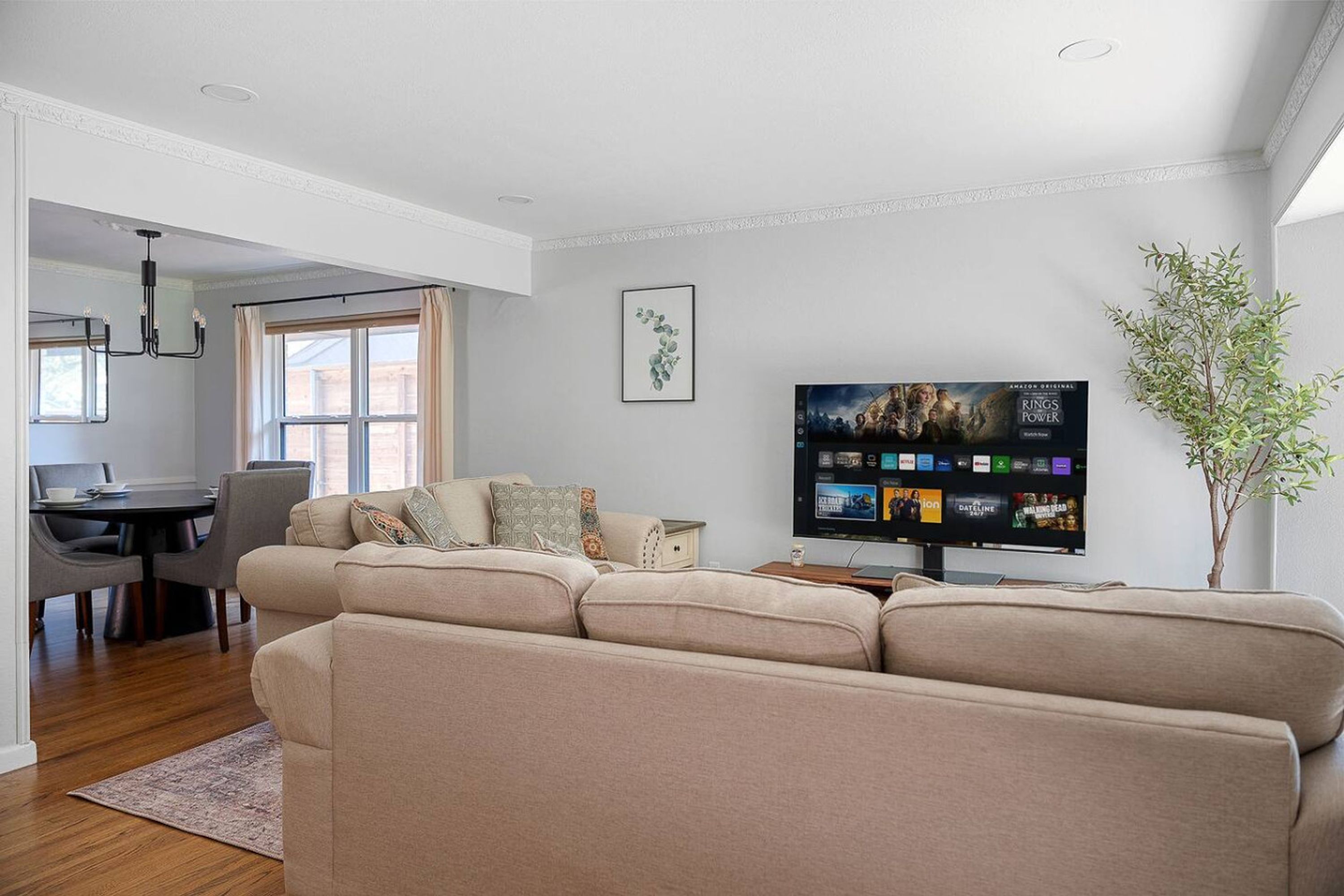 A living room with a beige sofa, a dining area with a table and chairs, a flat-screen TV mounted on the wall, and a potted plant.