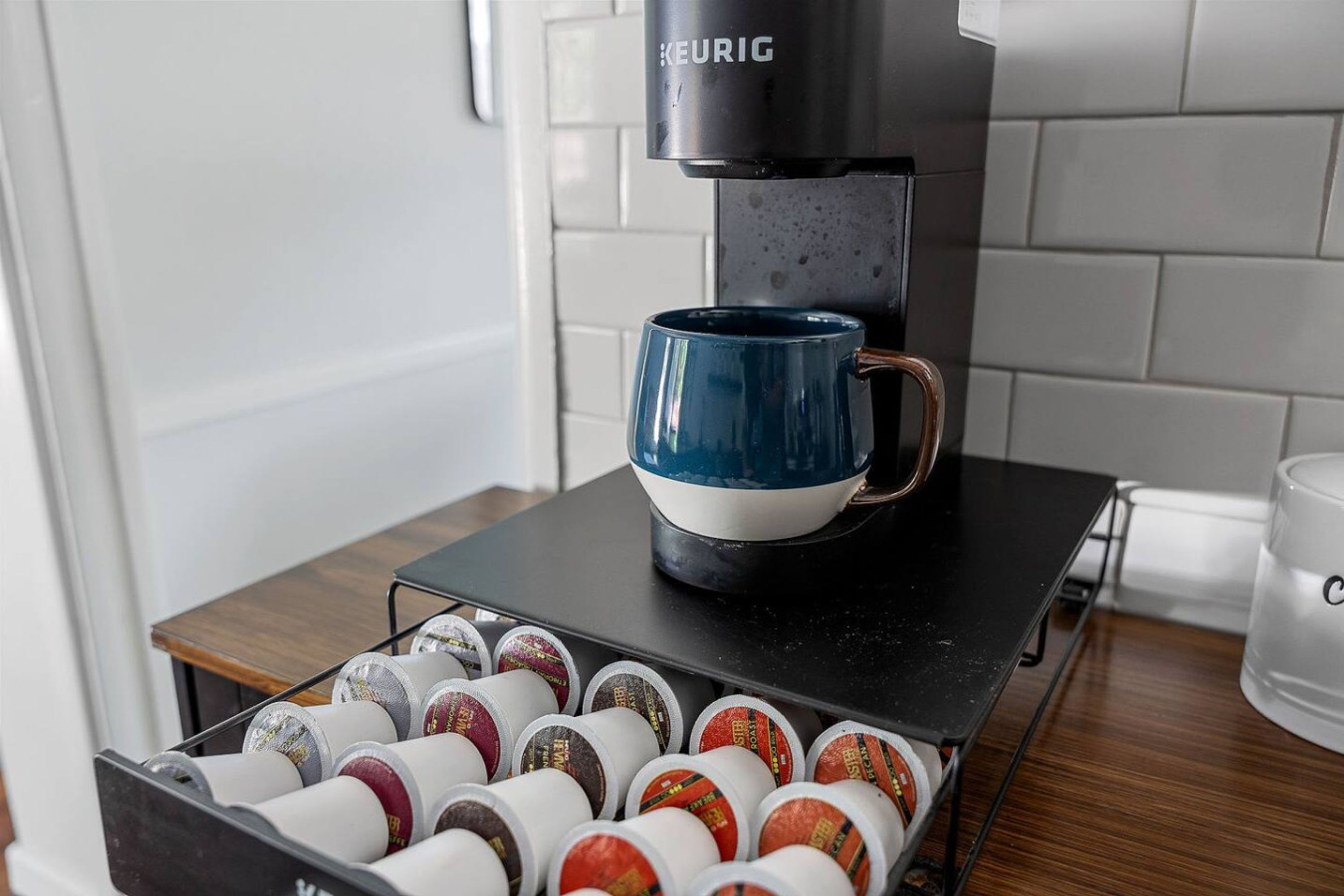 A Keurig coffee machine with a blue mug on the drip tray, a coffee pod holder with various pods, and a white mug on the counter.