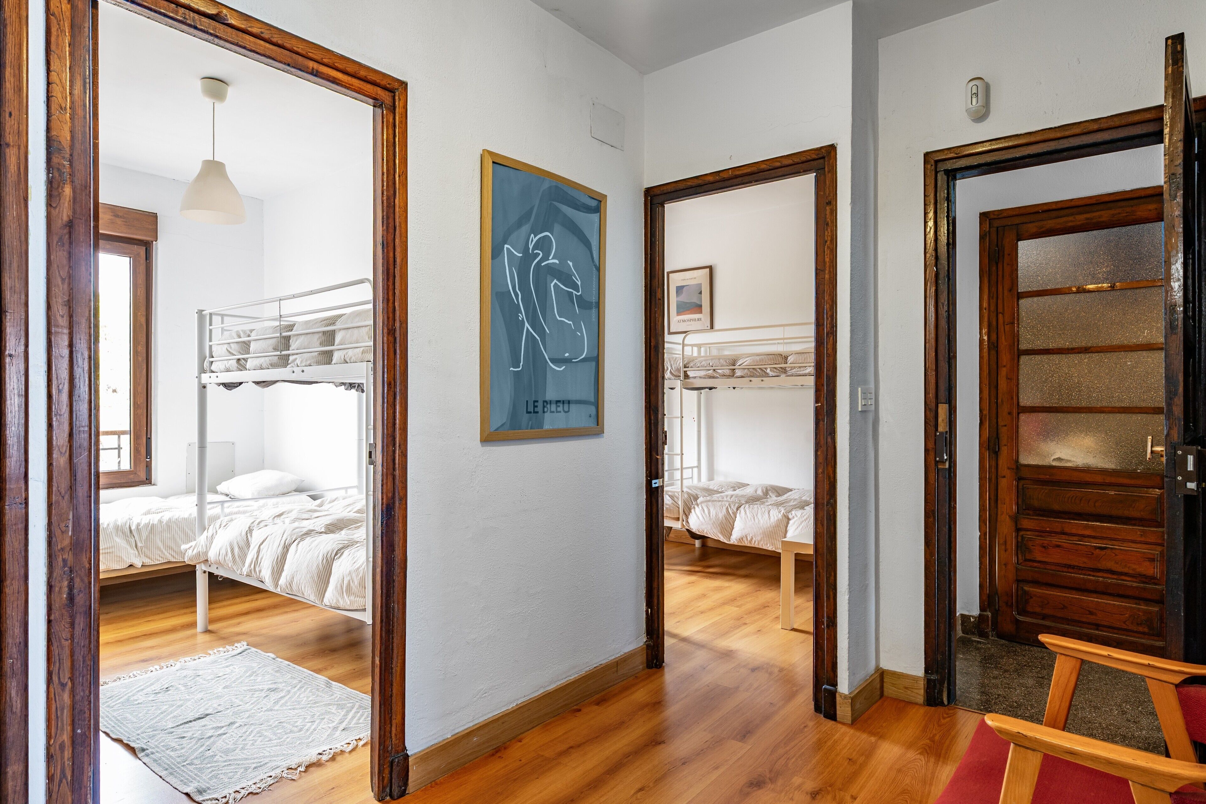 A room with two bunk beds, a wooden floor, a white wall with a framed picture, and a door with a glass panel.