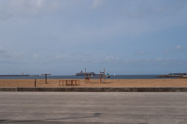 Ein Strand mit einer Betonpromenade, einer Holzkonstruktion und einem Schiff in der Ferne.