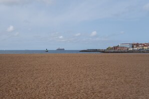 Ein weiter Sandstrand mit ruhiger See, ein Schiff in der Ferne und Gebäude auf der rechten Seite.