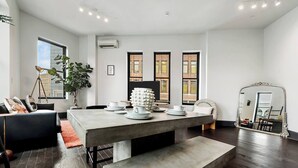 Un comedor moderno con una mesa grande, sillas y un banco. Hay dos ventanas con vistas a un edificio.
