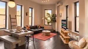 Una sala de estar moderna con un sofá, una mesa de centro y un televisor. Hay ventanas amplias y una planta.