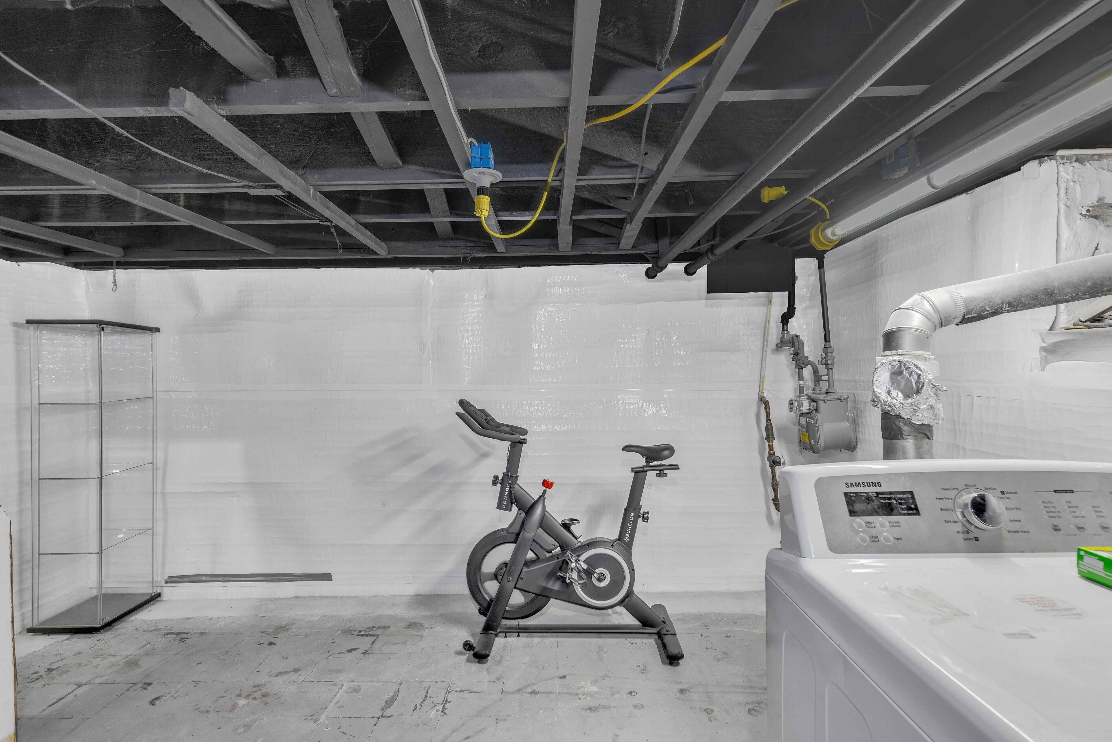 A room with a gray exercise bike, a white washing machine, and exposed ceiling wiring.
