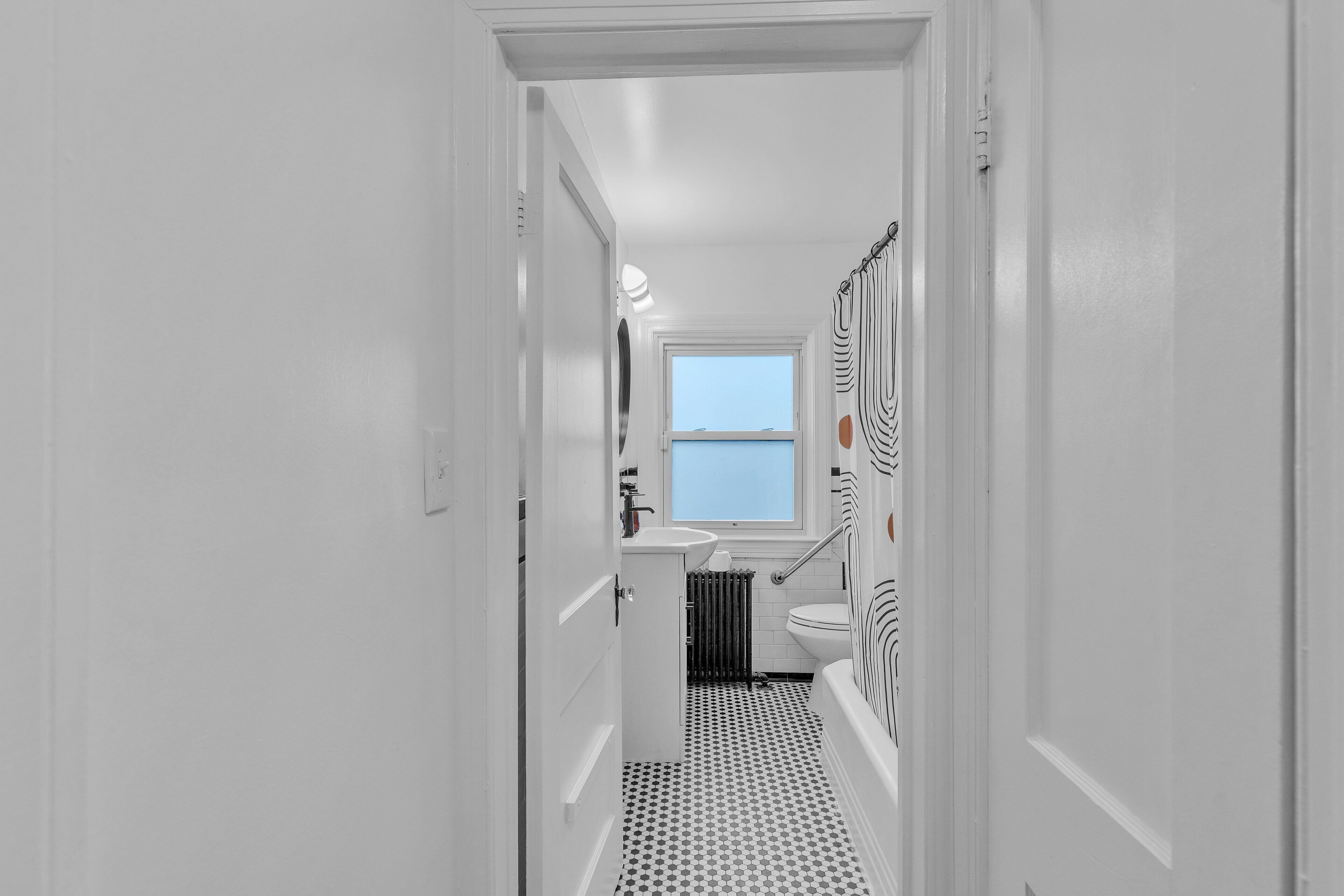 Een smalle gang die leidt naar een badkamer met een wit wastafelmeubel, toilet en bad.