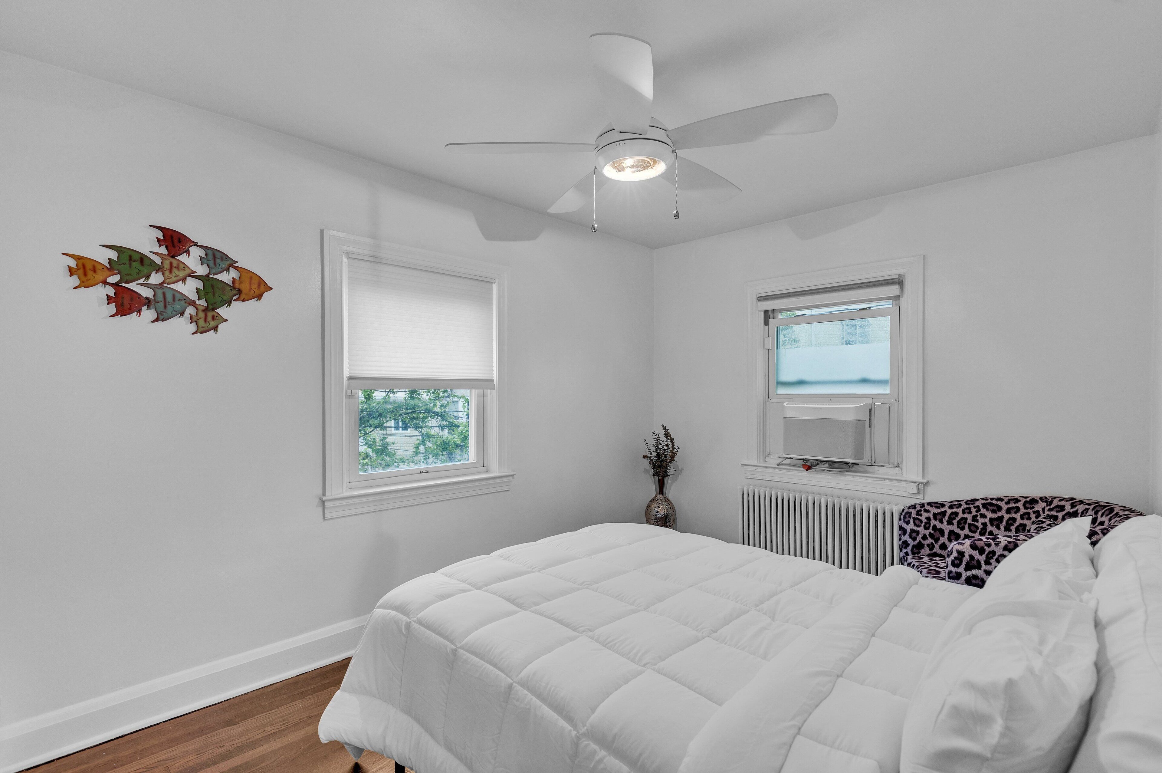 Une chambre avec un lit blanc, un ventilateur de plafond, une fenêtre avec des stores et un radiateur.