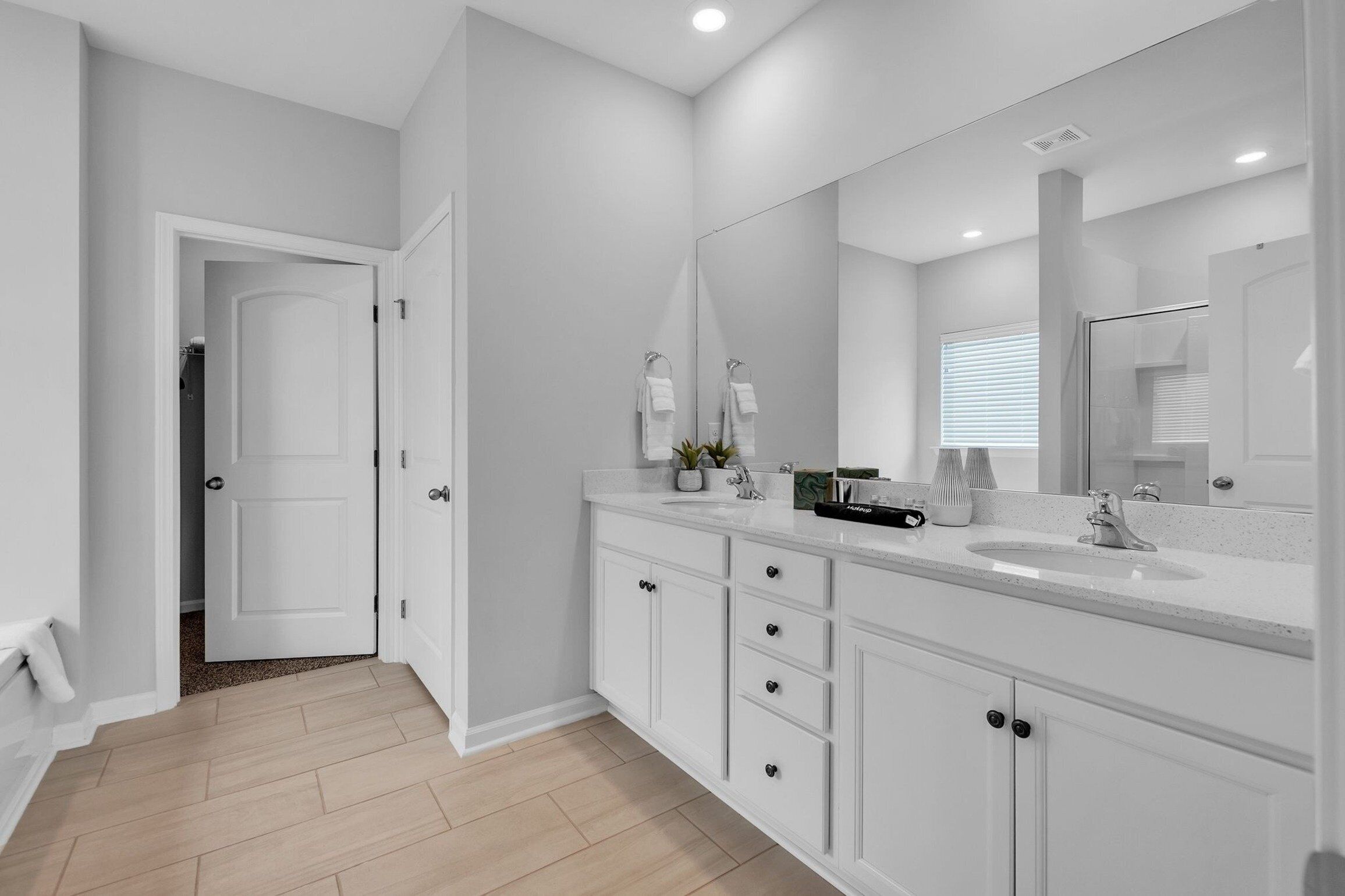 Une salle de bain moderne avec un grand miroir, des meubles blancs et un espace douche visible à travers une porte ouverte.