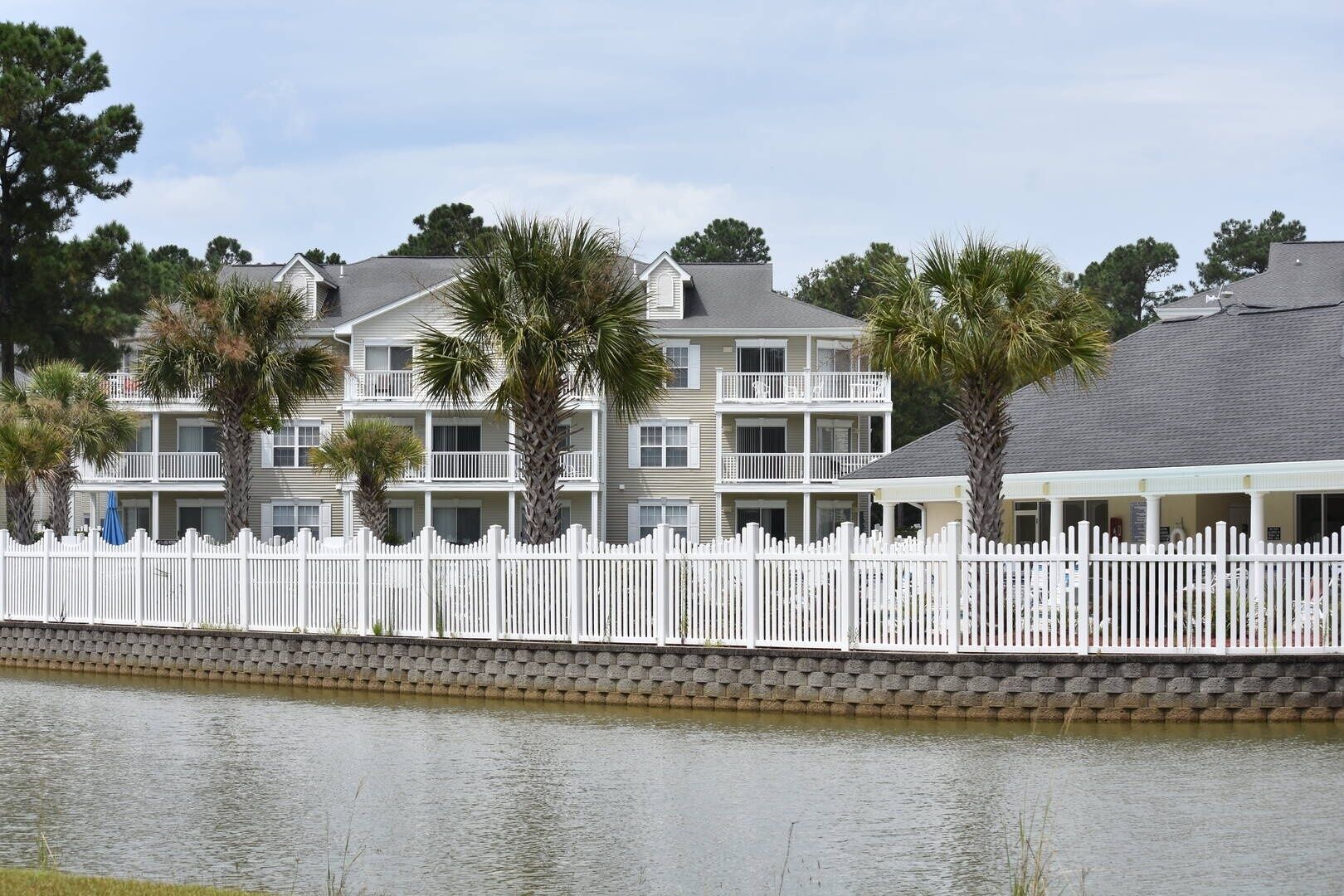 A waterfront hotel with multiple balconies, a white fence, and palm trees.