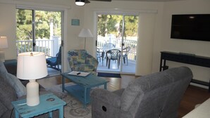 A living room with a ceiling fan, a TV mounted on the wall, a sofa, a chair, a small table with a lamp, and a view of the outside deck through large windows. - 2909 2 Bedroom Villa (Calabash)