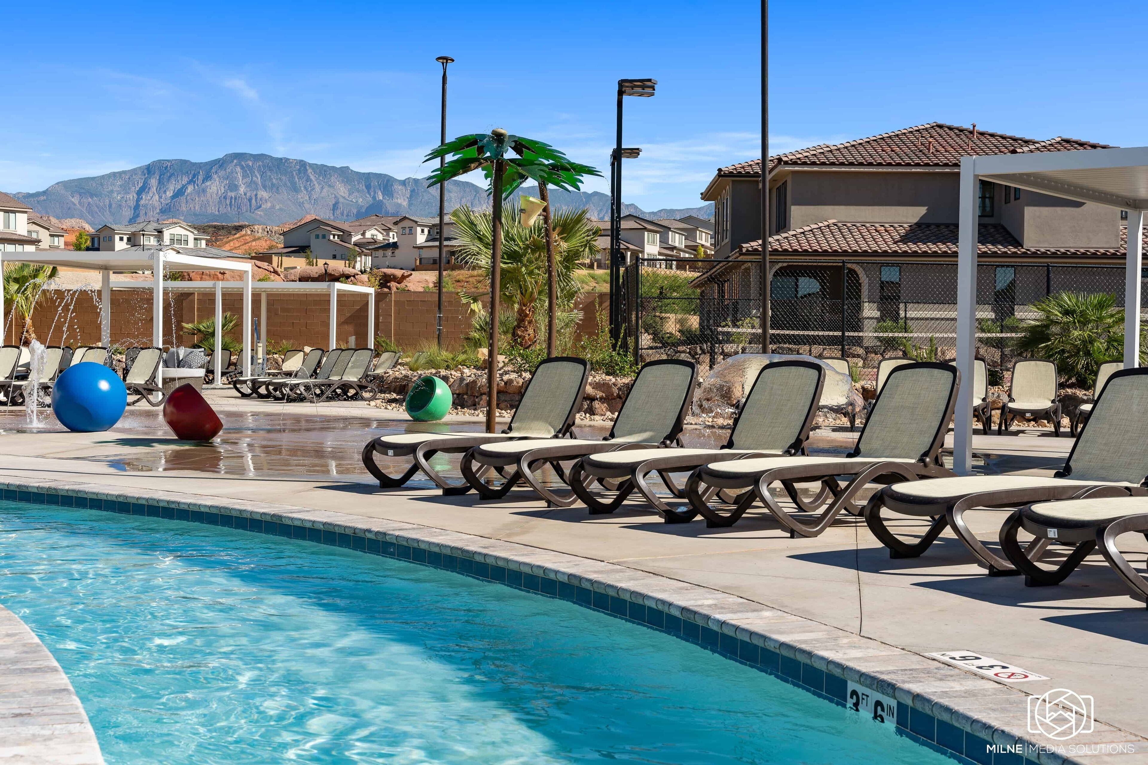 Ein Swimmingpool mit Liegestühlen und Übungsbällen, umgeben von einem Zaun und Palmen, mit Häusern und Bergen im Hintergrund.