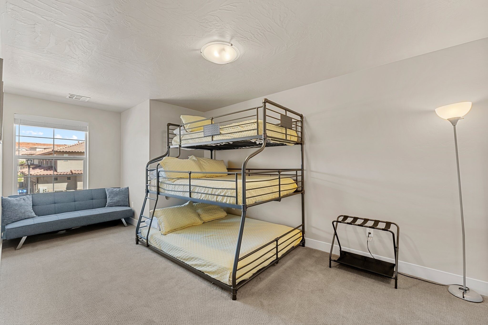 A bunk bed with two levels, a grey sofa, and a floor lamp in a room with a carpeted floor and a window.