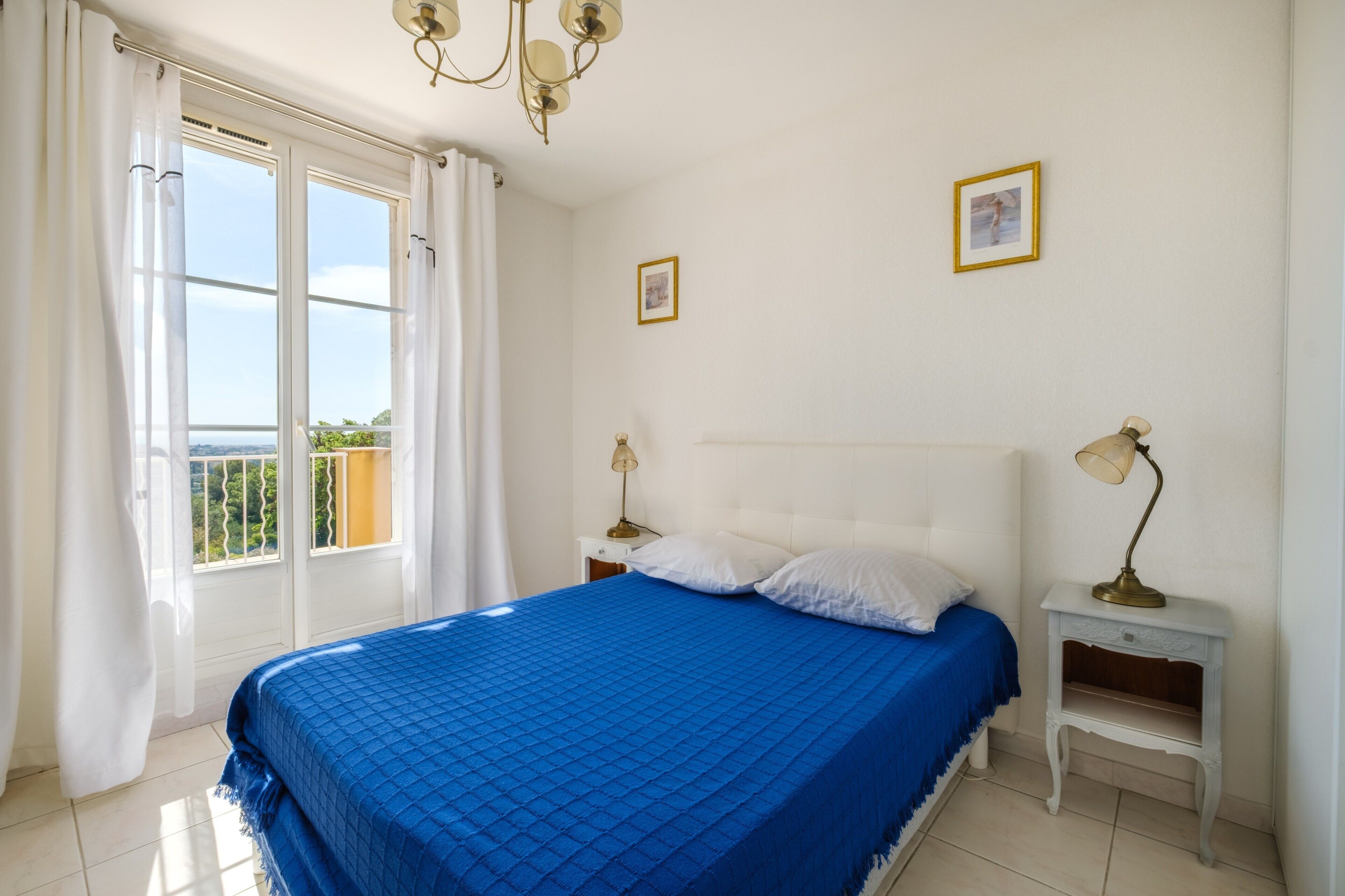 Een slaapkamer met een groot bed, twee nachtkastjes, een kroonluchter en uitzicht door het raam.