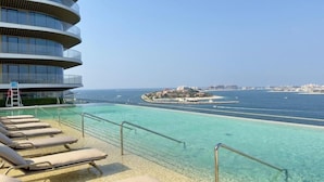 Ein Poolbereich mit Liegestühlen, freiem Blick auf das Meer und einem modernen Gebäude mit Balkonen.
