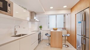 A modern kitchen with white cabinetry, stainless steel appliances, a central island with stools, and a window providing natural light. - Apartment 'Maina' with Sea View, Wi-Fi and Air Conditioning (Pollença)