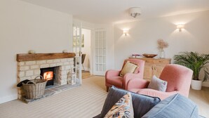 A cozy living room with a fireplace, a pink armchair, and a wooden cabinet. - Country Cottage in the Heart of the Cotswolds (Cheltenham)