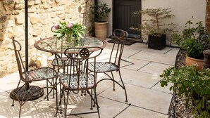 Une terrasse avec une table ronde en verre et des chaises en métal, entourée de plantes en pot et d’un mur en pierre.