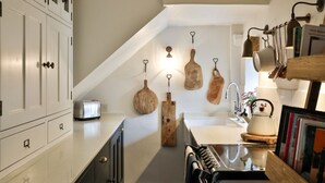 Une cuisine avec des meubles blancs, un grille-pain, des planches à découper en bois suspendues, une bouilloire et un évier.