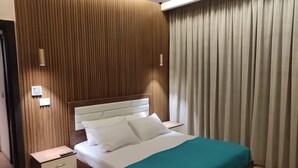 Camera d'albergo con parete rivestita in legno, un letto con biancheria da letto bianca e una coperta verde acqua, un comodino con telecomando e una finestra con tende.