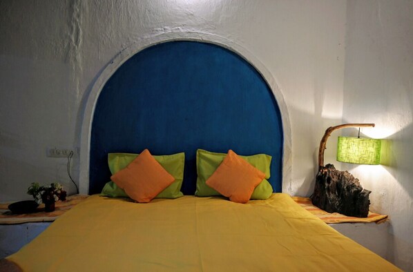 A bed with a yellow bedspread and two pillows, one green and one orange, in a room with a blue arched headboard and a wall-mounted lamp. - Bastar Birds Nest (Jagdalpur)