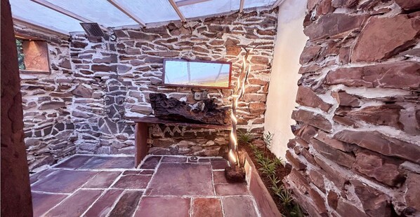 A stone-walled interior with a stone bench, a mounted television, and a fire feature. - Bastar Birds Nest (Jagdalpur)