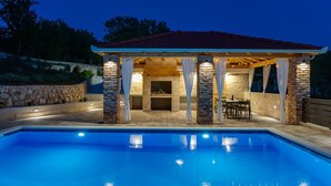 A covered outdoor seating area with stone pillars, a pool, and a stone wall in the background.