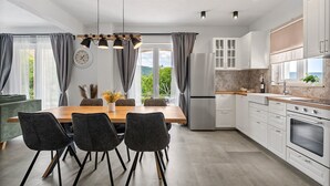 A modern kitchen with a wooden dining table, four chairs, a refrigerator, white cabinets, and a view of greenery outside.