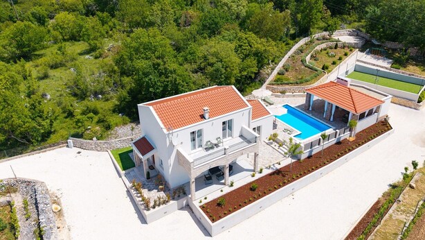 A modern two-story house with a swimming pool, surrounded by lush greenery and a stone retaining wall.